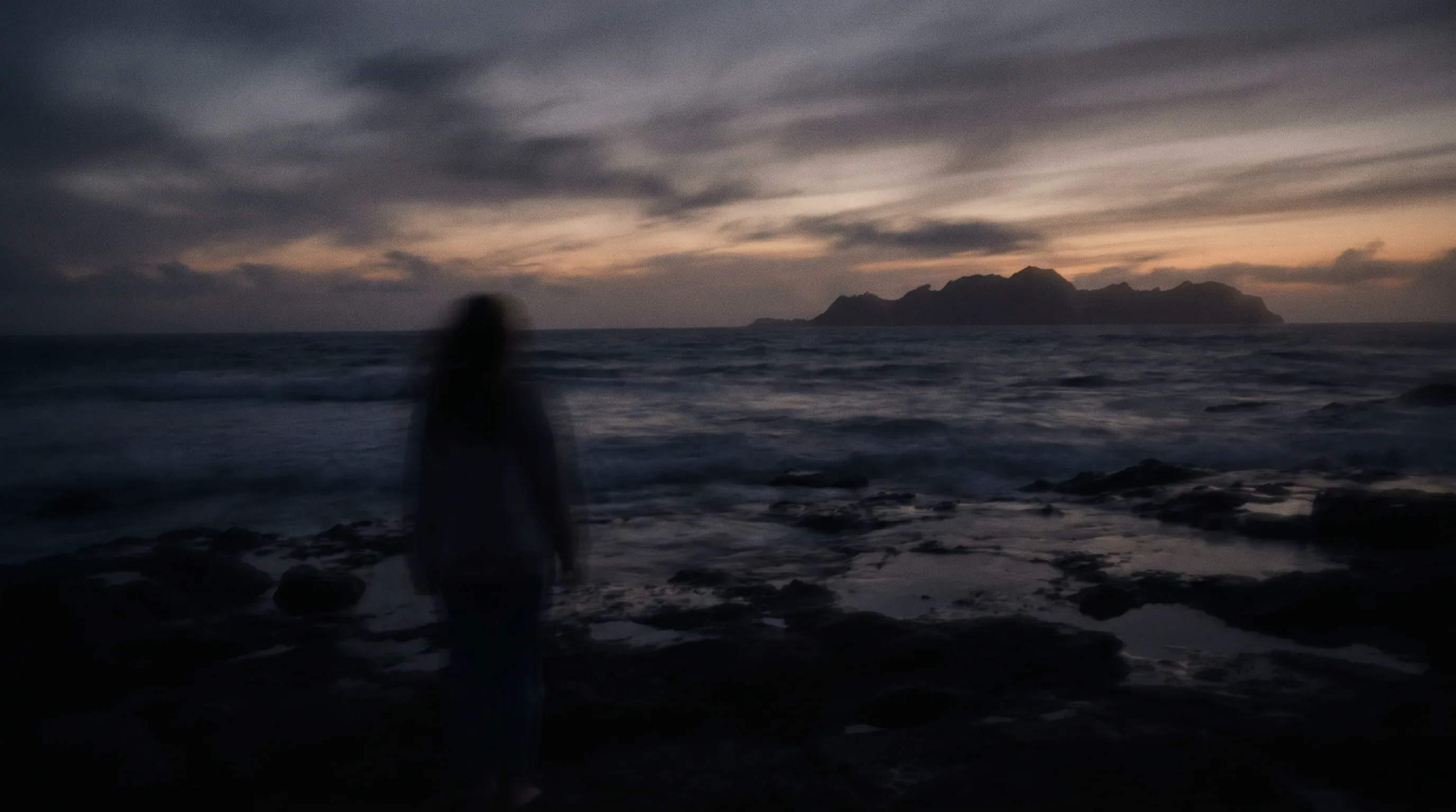 A blurred figure standing by the sea at dusk with an island on the horizon.